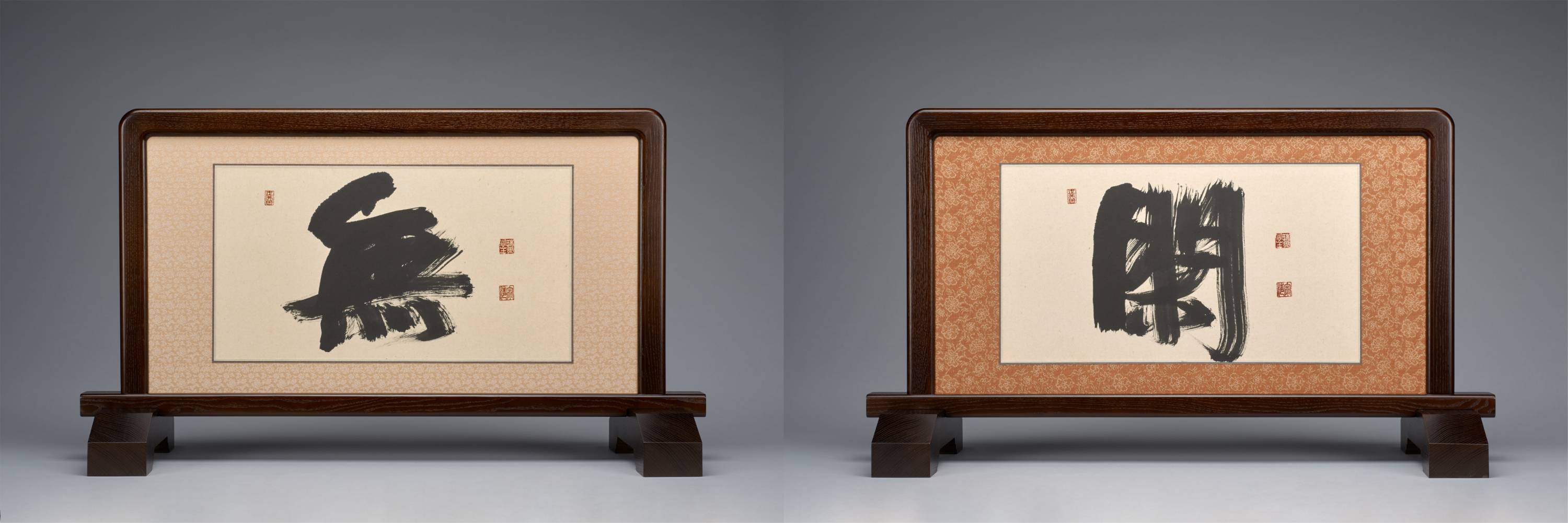 Small, double-sided table screen. Each face bears a sweeping, powerfully brushed Japanese character that occupies most of the paper ground. Small red seals punctuate it.