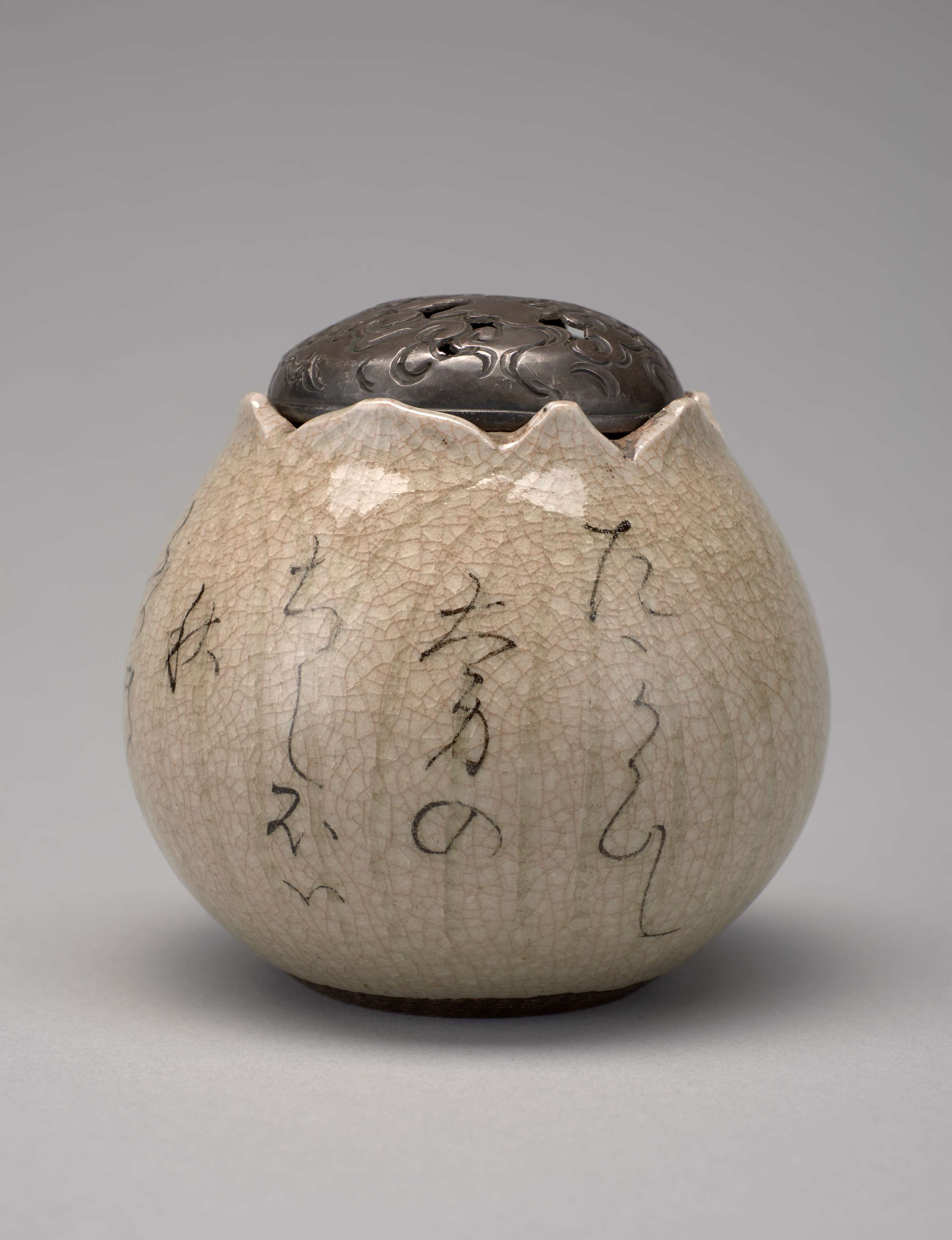 An incense burner with a round base that curves upward to an undulating rim. Thin Japanese calligraphy adorns the exterior. A darker silver cap sits atop the incense burner.