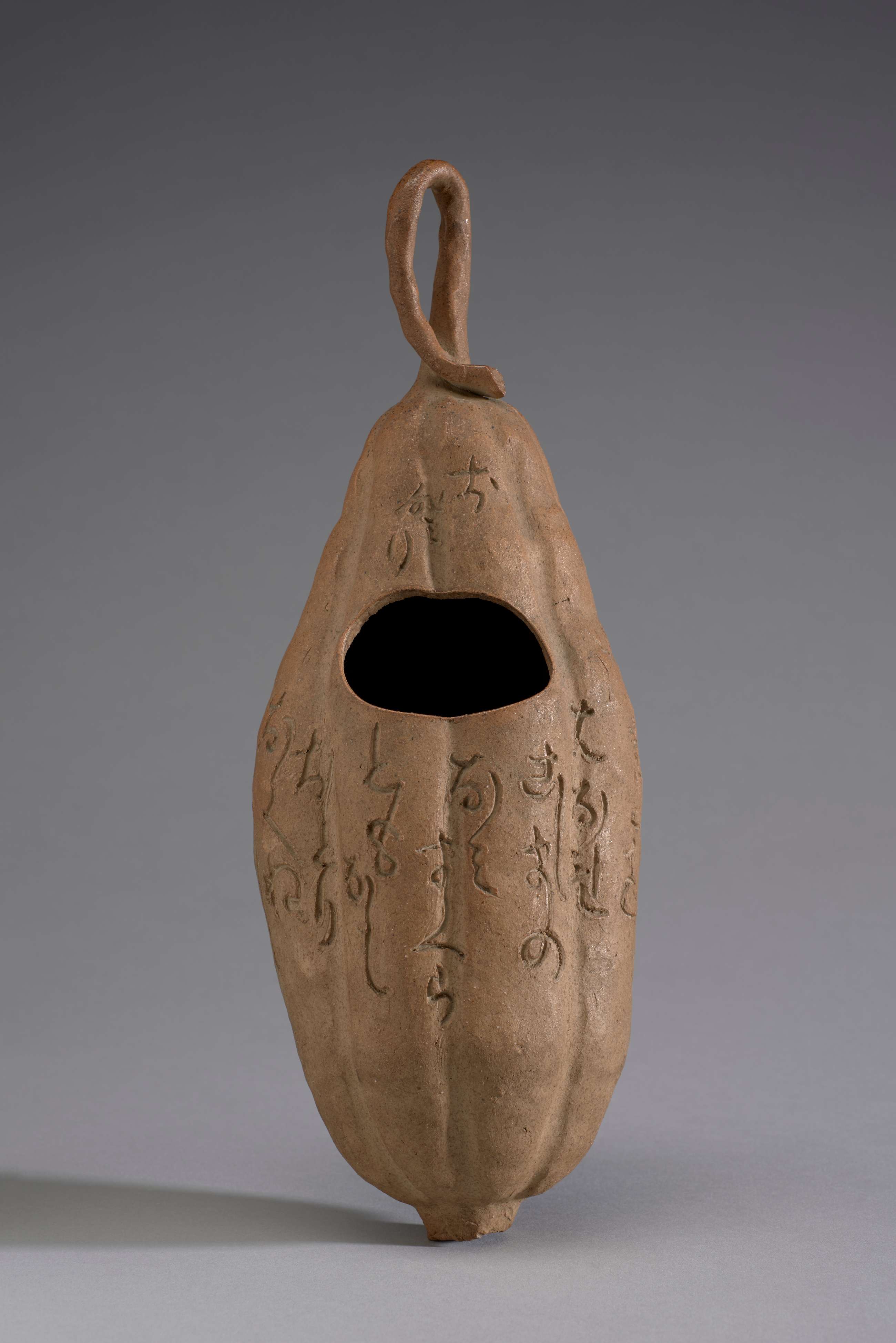 A gourd-shaped ceramic vase with vertical ridges and a loop of clay vine at the top. A dark hole near the center of the vase is surrounded by incised Japanese calligraphy.