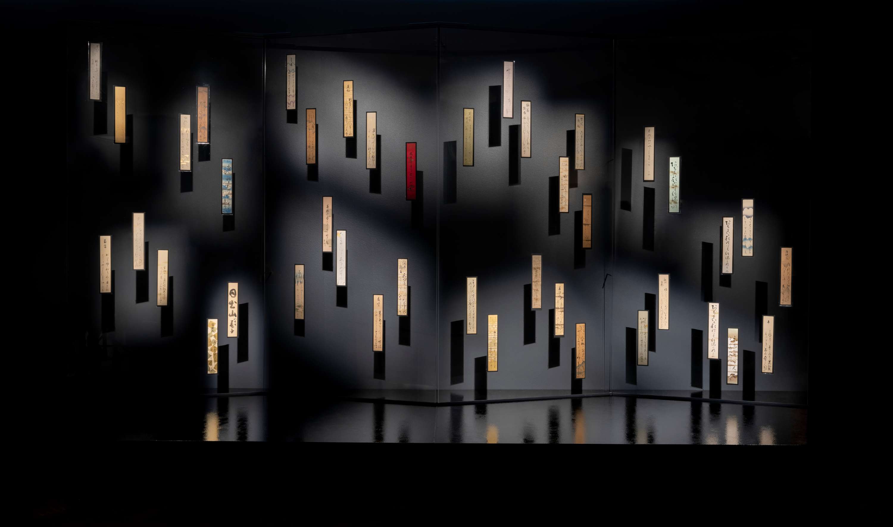 Dozens of vertical, decorated paper slips with calligraphy are mounted haphazardly on faux folding screen made of transparent plexiglass. Their reflection in the mirror-like platform, enhanced by the dark room, creates a sense of floating.