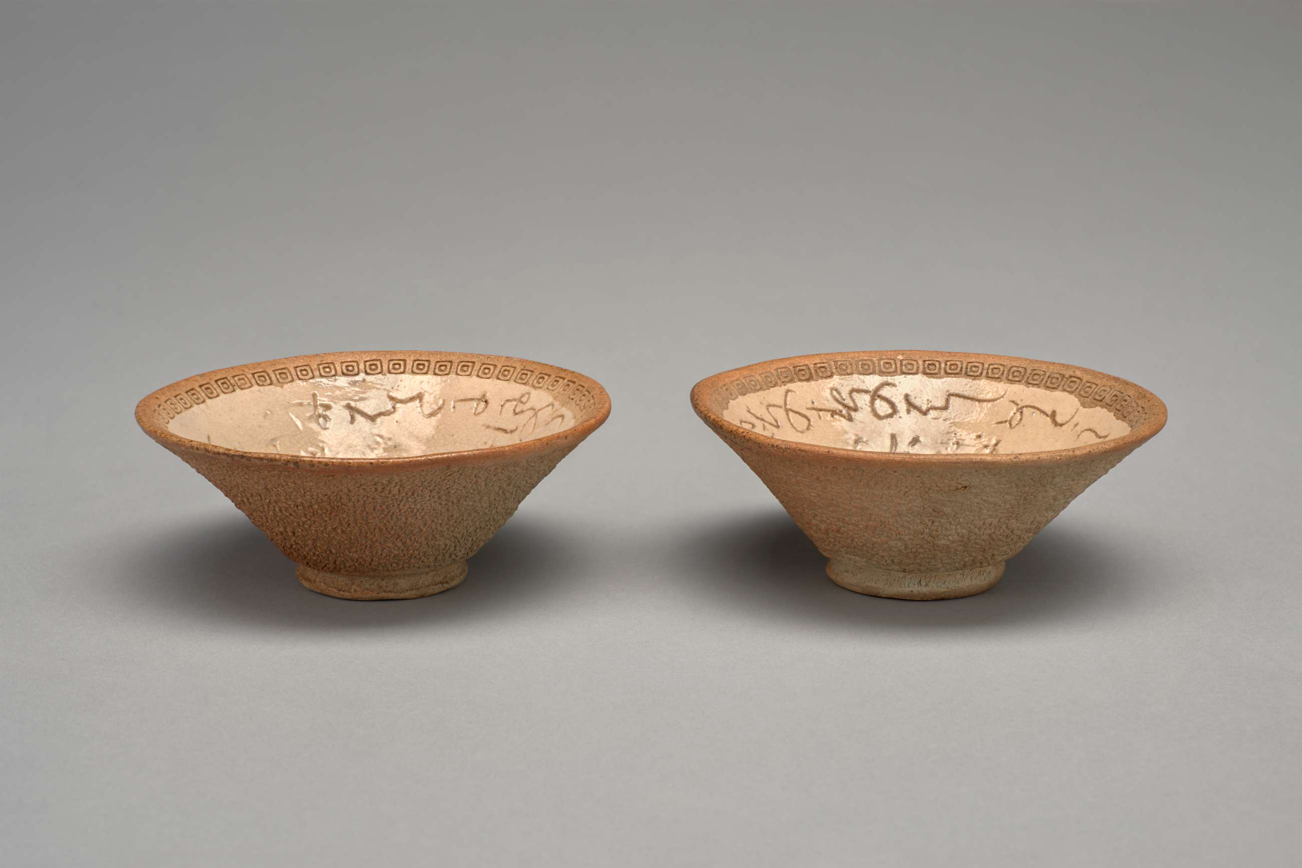 Two small beige bowls with slanted sides. The exteriors are rough and darker brown than the smooth interiors. The interior rims are stamped with small squares and circles. The interior of the bowls are lighter beige and inscribed with poems written in  Japanese characters.