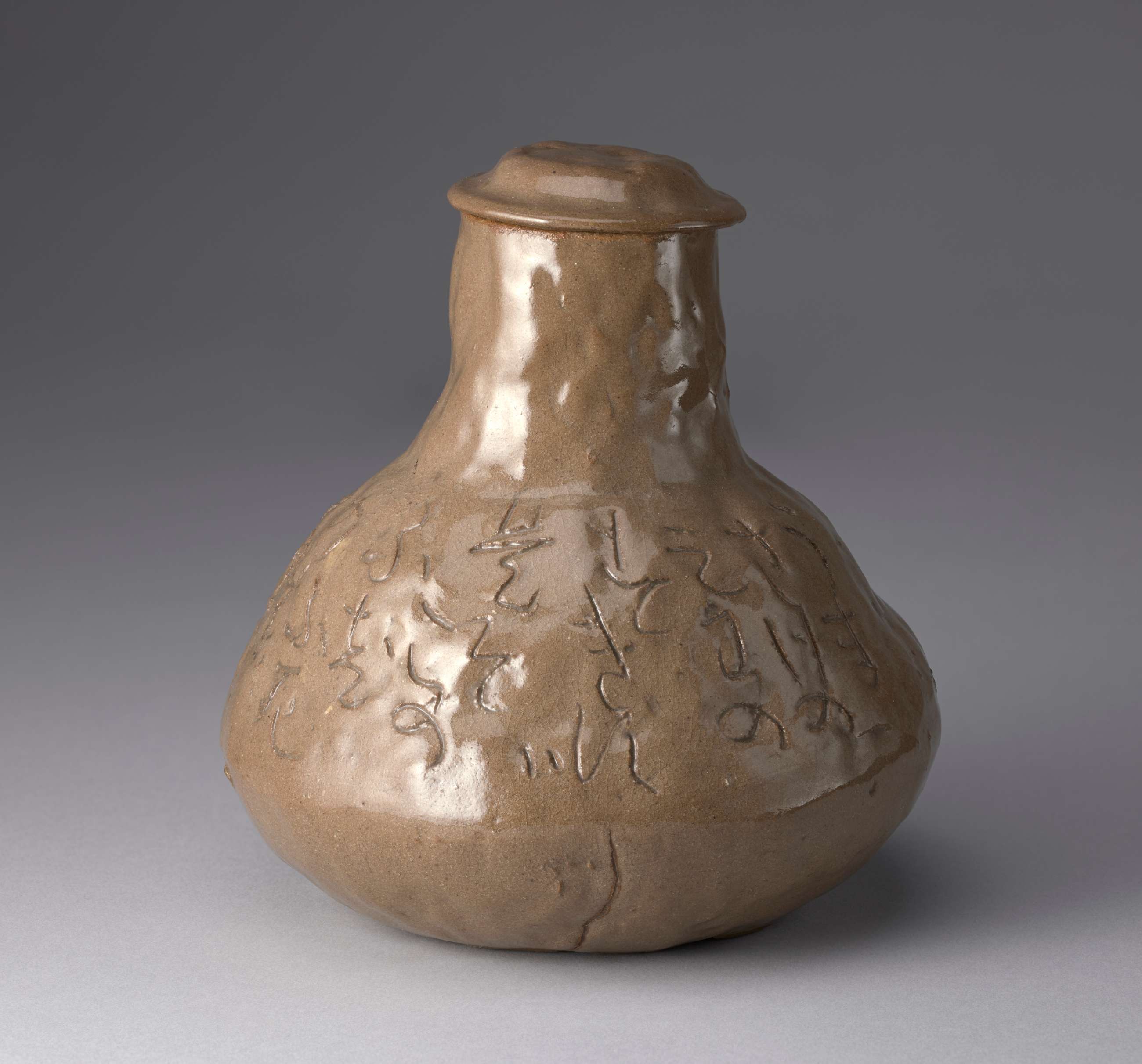 A light brown sake decanter with a narrow opening at the top and a much wider base. It has a small rimmed lid, and a poem in Japanese characters is inscribed around the vessel. Finger prints and indentations cover its surface.