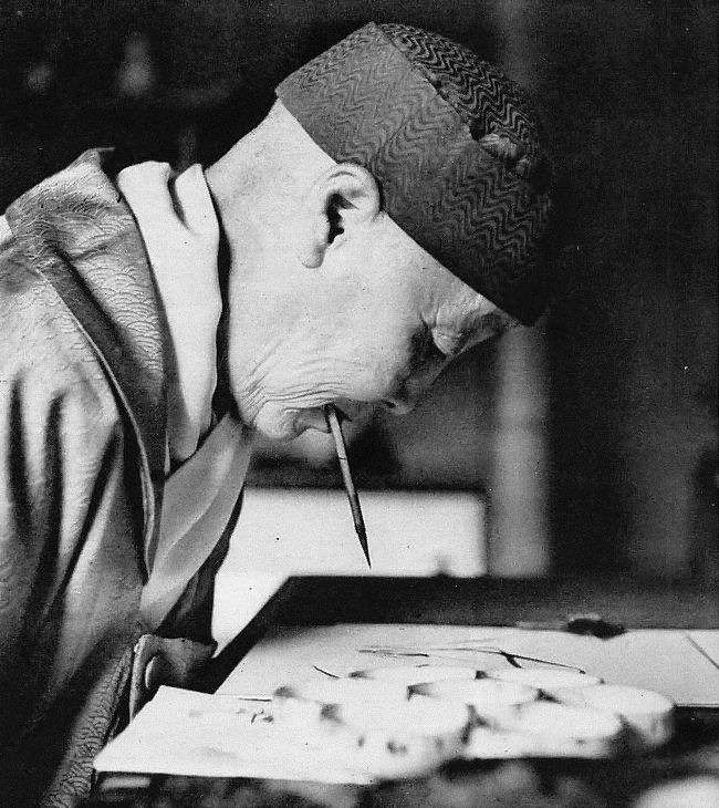 Black and white side-profile photograph of the artist wearing a black cap. She holds a paint brush in her mouth and bends towards a piece of paper on the table in front of her.