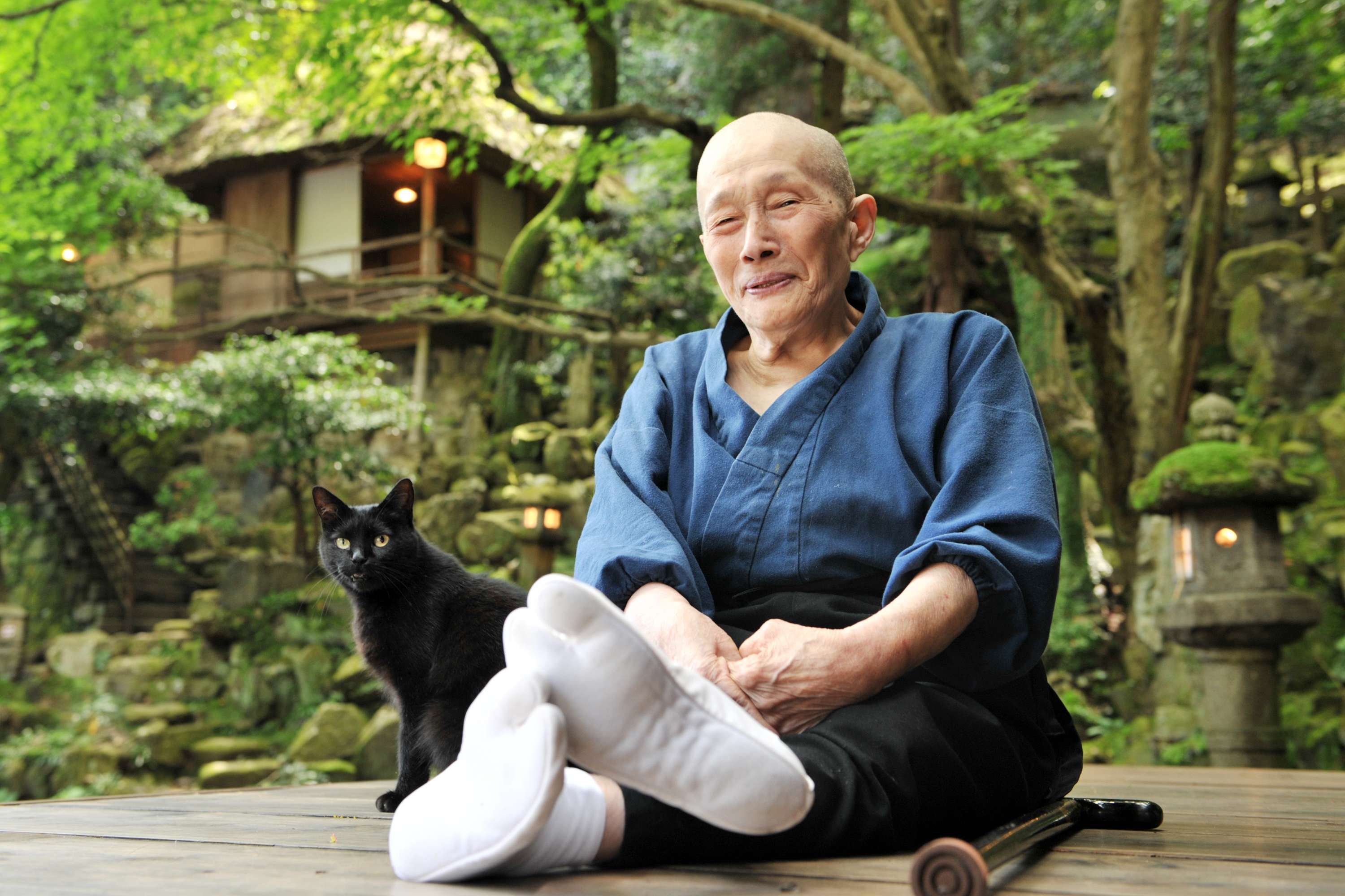 The nun-artist Murase Myōdō sits on a wooden platform, with her white-socked feet in the foreground. A black cat sits to the left, and a walking cane lies on the platform on the right. A lush green forest and a thatch-roofed house in the background.