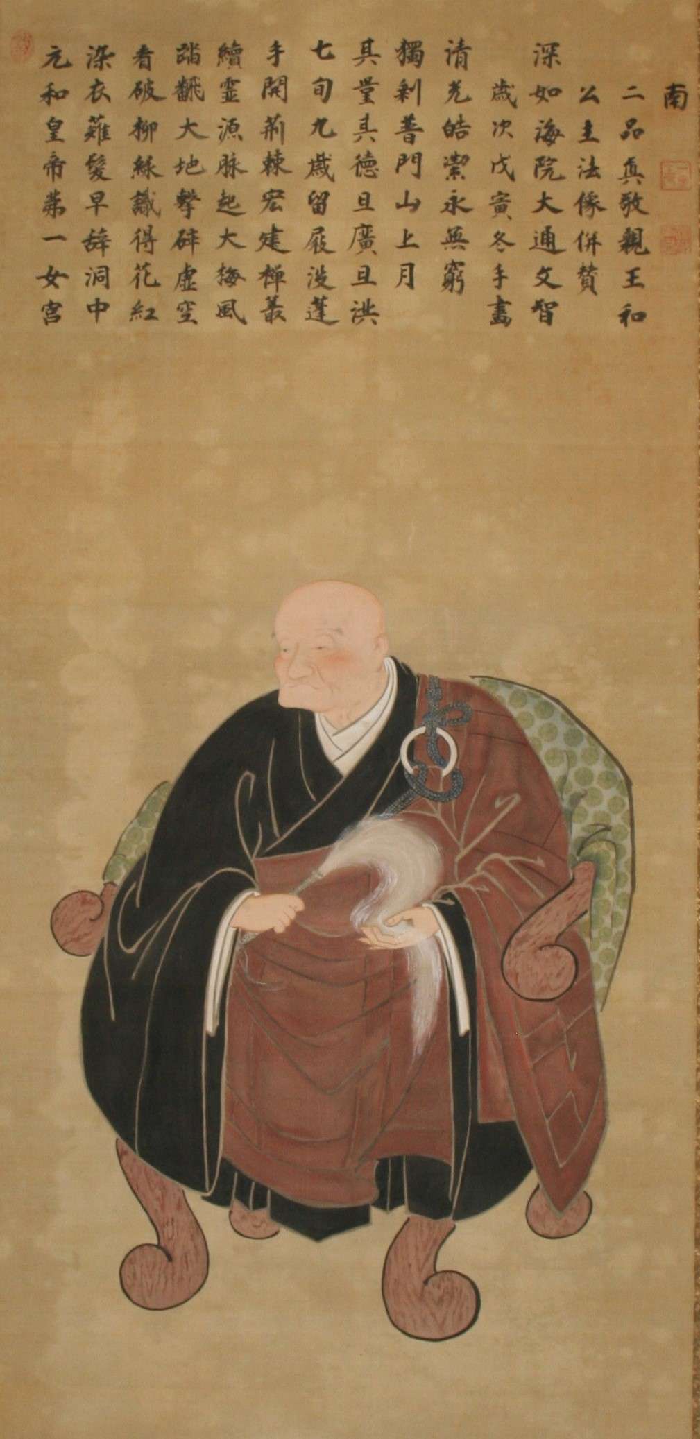 An older woman with no hair wears heavy black and brown robes and sits on a green polka-dot armchair with wooden arms and legs. The shape of the chair matches the woman’s round body. Above her, fourteen lines of Japanese characters fill the top of the frame.