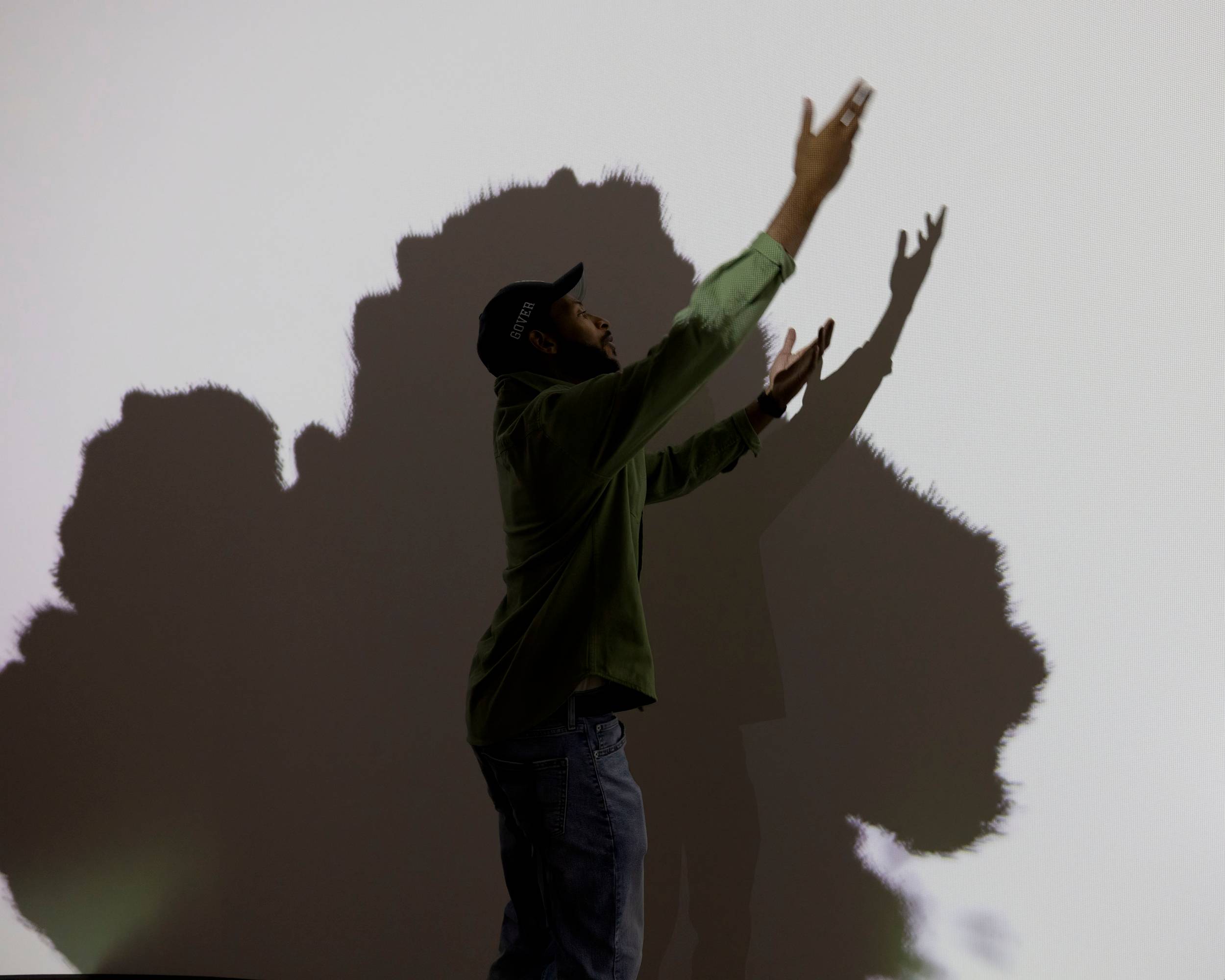 A person stands in the center, look up and to the right with arms outstretched. Behind, a black inky splotch fills a white wall.