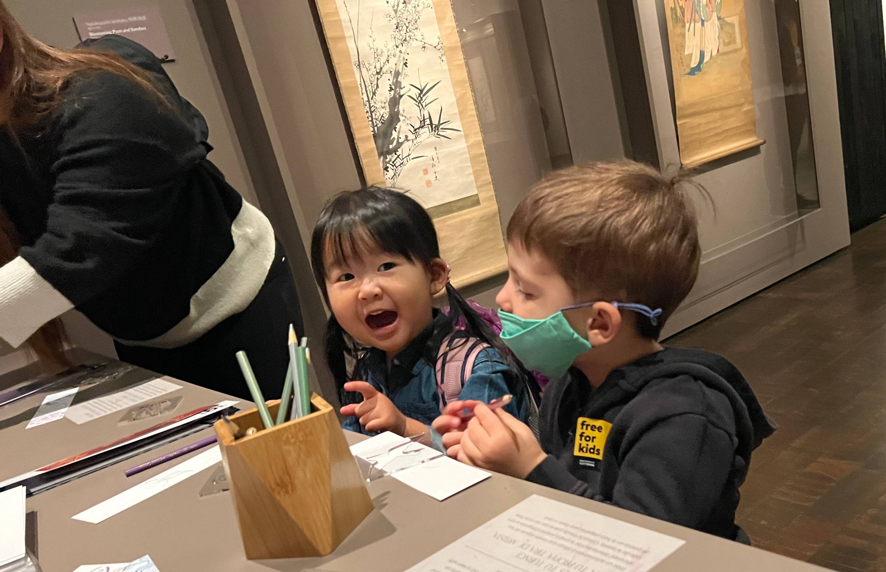 A young child looks at the camera with mouth open. Another child wearing a mask looks at the paper on a long table.